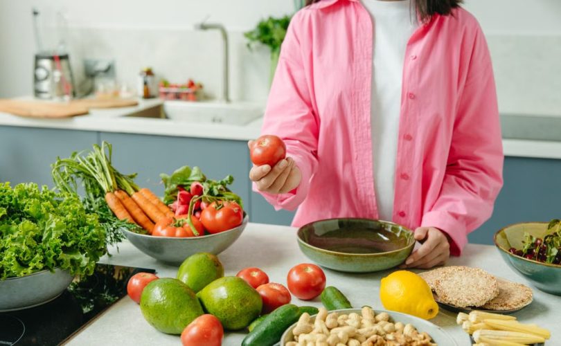 Woman in pink overshirt holds tomato in kitchen with fresh fruits and veggies.